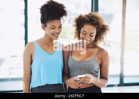 Raccontiamo a tutti la nostra sessione di allenamento. Due giovani donne in forma che chiacchierano mentre guardano un telefono all'interno di una palestra prima di una sessione di allenamento. Foto Stock