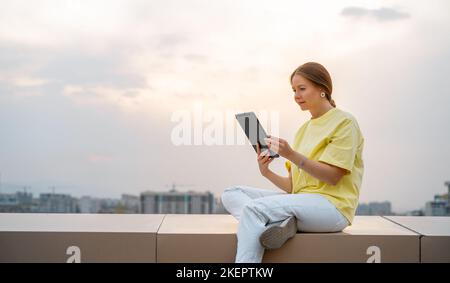 La giovane donna lavora su un tablet pc seduto sul tetto o sulla terrazza del grattacielo in estate la mattina del giorno. Foto Stock