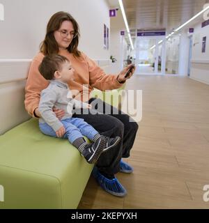 Madre e bambino con un telefono cellulare in clinica in attesa di un pediatra. Genitore con un bambino con uno smartphone nelle mani. Il bambino è un ragazzo Foto Stock