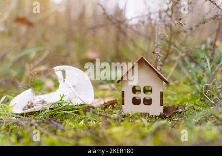 Concetto di ecologia. C'è una piccola casa di legno sull'erba, accanto ad essa c'è un vetro di plastica - rifiuti. Foto Stock