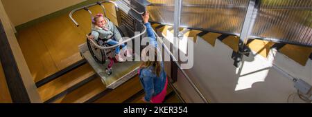 Madre con un bambino che vive con paralisi cerebrale utilizzando un ascensore elettrico per sedie a rotelle per accedere all'edificio pubblico. Speciale piattaforma di sollevamento per sedia a rotelle Foto Stock