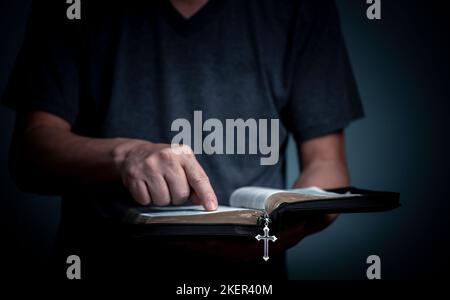 Giovane che legge la Sacra Bibbia e indica la parola con la collana a croce in font del libro. Foto Stock