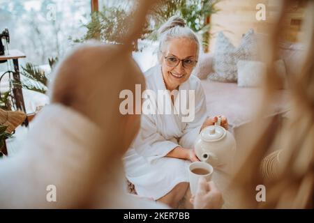 Coppia senior in accappatoi che si gode il tempo insieme nel loro soggiorno, bevendo tè caldo, calma e l'atmosfera hygge. Foto Stock