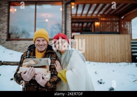 Portait di coppia anziana che trasporta i tronchi di legno e che prepara il fuoco all'aperto, durante il giorno di inverno. Foto Stock