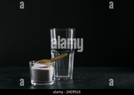 Polvere di collagene in recipiente, bicchiere d'acqua e misurino su fondo nero Foto Stock