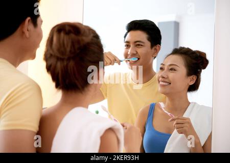 Giovane coppia asiatica allegra guardando lo specchio e spazzolando i denti insieme Foto Stock