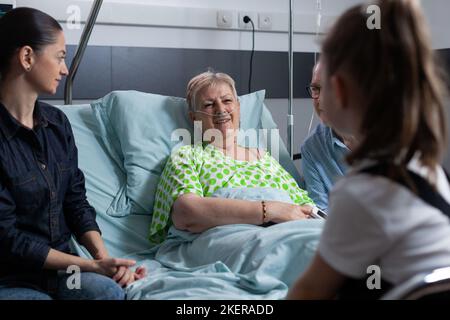 Donna anziana sorridente sotto osservazione medica in ospedale, chiacchierando felicemente con la ragazza. Parenti che visitano la signora anziana durante il soggiorno in ospedale. Felice paziente anziana con diverse persone in sala sanatoria. Foto Stock