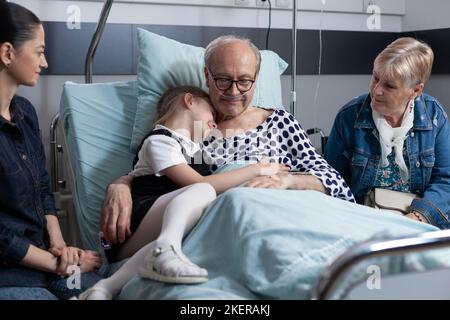 L'uomo anziano nella camera da letto della clinica geriatrica abbraccia la bambina. Parenti anziani che visitano la sala di ricovero della torre medica. Nipote visita malato nonno al sanatorio. Foto Stock