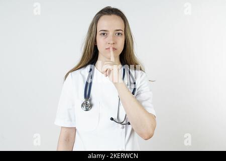 Medicina e concetto di salute. Una giovane donna medico mostra con un gesto del dito che il silenzio dovrebbe essere osservato. Isolato su sfondo bianco. Foto Stock