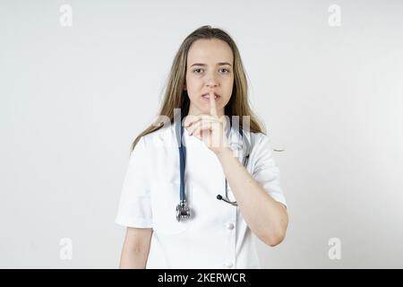 Medicina e concetto di salute. Una giovane donna medico mostra con un gesto del dito che il silenzio dovrebbe essere osservato. Isolato su sfondo bianco. Foto Stock