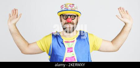 Concetto di vacanza. Clown - un uomo in un vestito blu e giallo brillante, occhiali e un cappello emotivamente sollevato le mani. Foto Stock