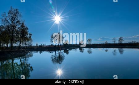Paesaggio di lago con riflesso del sole che è su cielo blu, Silhouette di alberi sullo sfondo Foto Stock