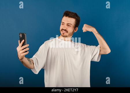 Ritratto dell'uomo allegro che indossa una t-shirt bianca in posa isolata su sfondo blu prendendo selfie, mostrando braccio forte, flettendo bicipiti e posando per p Foto Stock