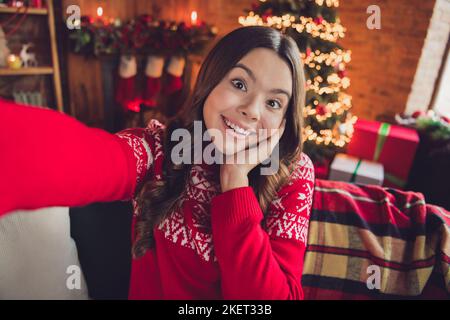 Ritratto di ragazza ecstatica impressionata con capelli ondulati vestito ornamento rosso maglione facendo selfie palma su zigomi in casa al coperto Foto Stock