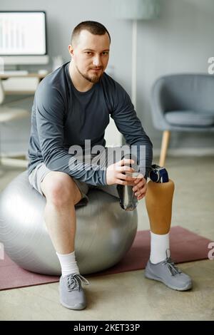 Ritratto verticale completo dell'uomo con gamba protesica seduta sulla palla fitness e tenendo una bottiglia d'acqua dopo l'allenamento a casa Foto Stock