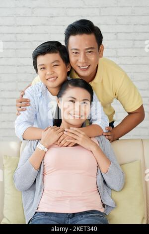 L'amorevole famiglia asiatica di tre persone si è riunita in un accogliente salotto e si è posata per fotografare con sorrisi ampi, foto di gruppo Foto Stock