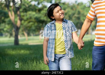 Ritratto di carino ragazzino in casualwear tenendo le mani con il papà e guardandolo con ampio sorriso mentre godendo piacevole passeggiata al parco verde illuminato da raggi di sole Foto Stock