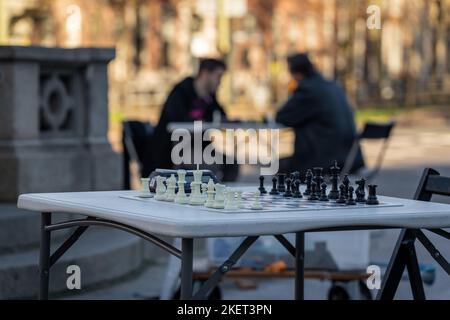 Scacchiera per strada pronta per una partita. Silhouette di giocatori di scacchi al tavolo sullo sfondo sfocato. Foto Stock