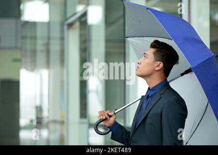 Vista del profilo di un giovane imprenditore asiatico che indossa un abito elegante tenendo in mano un ombrello e guardando verso l'alto, all'interno di un moderno edificio di uffici sullo sfondo Foto Stock