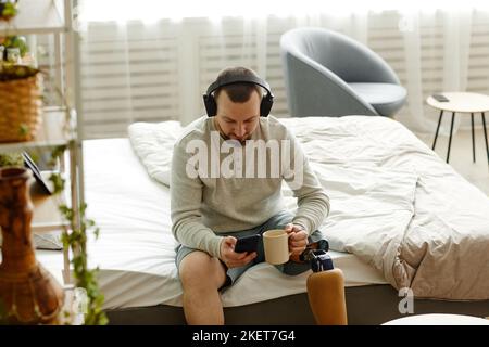 Ritratto ad angolo alto dell'uomo con gamba protesica che si gode la mattina a casa e l'ascolto di musica con cuffie, spazio copia Foto Stock