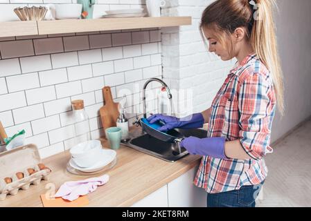 Giovane donna che indossa guanti di gomma lavando padella in cucina. Foto Stock