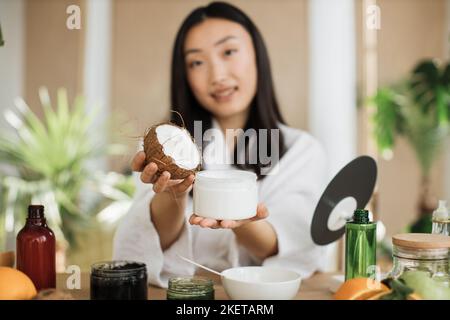 Focus su vaso di cocco e vetro, giovane donna asiatica che tiene metà di cocco e fare crema fatta in casa per la pelle sana. Molti ingredienti per cosmetici fatti in casa sdraiati su un tavolo di legno. Foto Stock