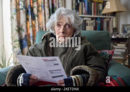 Ritratto della donna anziano che indossa il cappotto al coperto cercando di mantenere caldo a casa in crisi energetica guardando la bolletta energetica Foto Stock