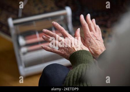 Primo piano di Donne anziane riscaldando mani su riscaldatore elettrico in cosyt di crisi di energia vivente Foto Stock