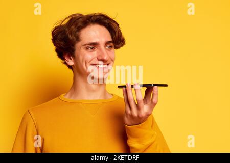 Ritratto di un giovane uomo con capelli ricci in posa, registrazione di messaggi vocali al telefono isolato su sfondo giallo Foto Stock