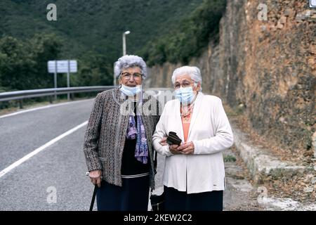 due anziane donne caucasiche con capelli bianchi e occhiali che camminano sulla strada pericolosa accanto alla foresta sorridendo guardando la macchina fotografica con maschera facciale Foto Stock