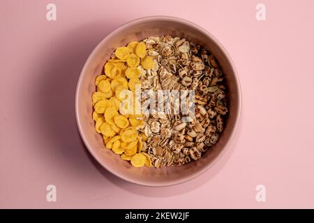 Vari tipi di cereali per la colazione in una ciotola rosa. Fiocchi di mais avena arrotolata e frumento soffiato. Sfondo rosa. Foto Stock