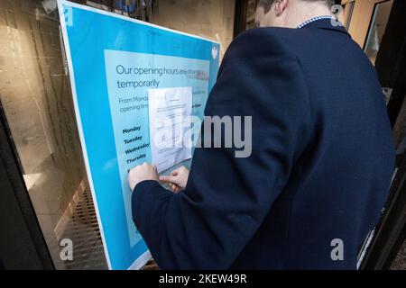 Un membro dello staff della Barclays Bank inserisce un avviso per i clienti all'ingresso della propria filiale Castle Place a Belfast, avvisando che al momento non possono aprirsi a causa di circostanze che esulano dal loro controllo. Due persone sono state arrestate per sospetto di danni criminali e comportamento disordinato dopo che la vernice rossa è stata gettata sopra la banca durante una piccola protesta. Data immagine: Lunedì 14 2022 novembre. Foto Stock