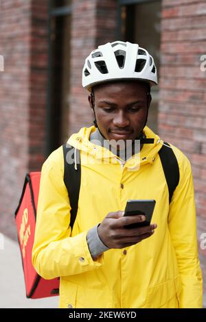 Giovane uomo nero in casco e giacca gialla guardando lo schermo del suo smartphone mentre consegna cibo ai clienti di società di servizi online Foto Stock
