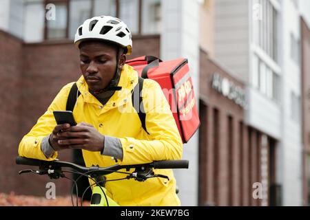 Serio uomo nero in casco di sicurezza e uniforme di lavoratore di servizio di consegna di cibo seduto in bicicletta e controllare nuovi ordini online al telefono Foto Stock