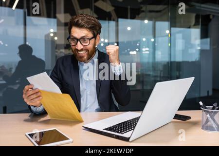 Uomo d'affari maturo felice con le buone notizie ha ricevuto la lettera nella busta, uomo anziano con la barba che legge e sorridente che lavora all'interno dell'ufficio al lavoro usando il laptop, l'investitore che celebra la vittoria di conquista. Foto Stock