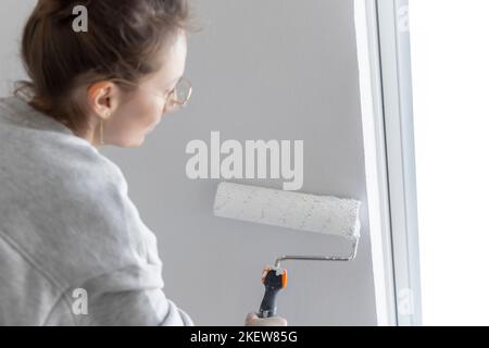 Vista ravvicinata giovane donna adulta pittura a mano muro pittura tenendo rullo pennello bianco colore soggiorno in movimento nuovo appartamento casa . Pittura femminile decorazione Foto Stock