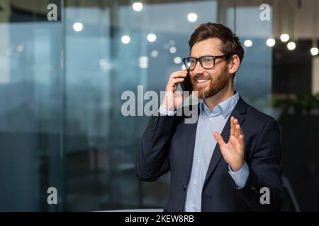 Uomo d'affari maturo in una tuta d'affari che parla al telefono, il manager è soddisfatto del risultato del risultato e il lavoro parla del risultato positivo, l'uomo è al lavoro all'interno dell'ufficio. Foto Stock