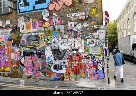 un vivace muro di mattoni coperto di graffiti e arte di strada incollata su Buxton Street appena fuori Brick Lane nell'est di Londra, Regno Unito Foto Stock