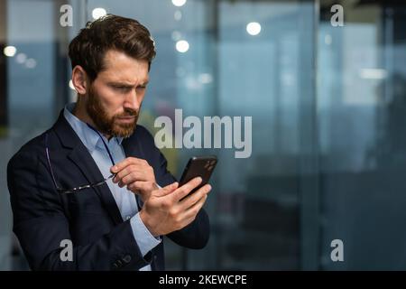 Uomo d'affari maturo in ufficio vicino alla finestra di sera sconvolto la lettura messaggio on-line di notifica dal telefono, capo maschio che lavora all'interno dell'ufficio in tuta d'affari e occhiali con barba, sconvolto investitore. Foto Stock