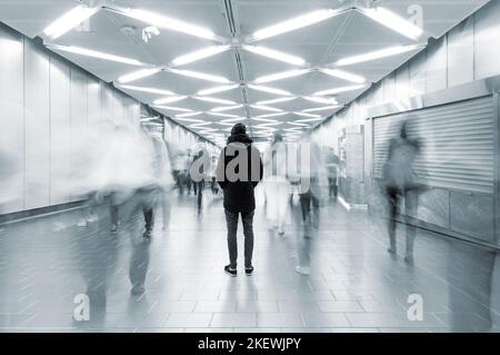 Foto a lunga esposizione con un uomo solitario girato da dietro in una passerella trafficata con persone che camminano in movimento sfocate, concetto di vita cittadina affollata Foto Stock