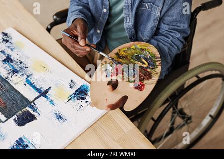 Vista dall'alto di un giovane uomo nero con tavolozza di colori che mescola vernici acriliche mentre si siede su sedia a rotelle da un tavolo di legno con tela Foto Stock