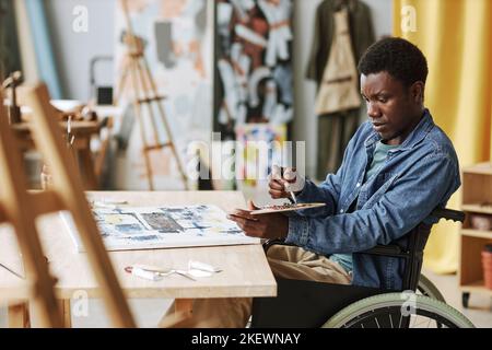 Giovane artigiano afroamericano in sedia a rotelle che mescola i colori sulla tavolozza mentre si siede al tavolo in studio di arte e pittura su tela Foto Stock
