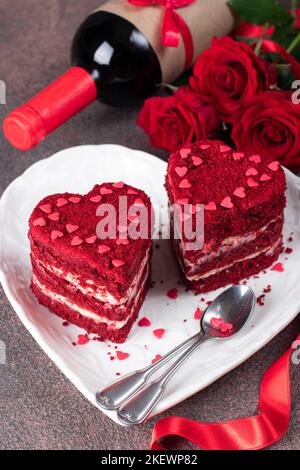 Torte velluto rosso a forma di cuori su piatto bianco, rose e vino per San Valentino su sfondo marrone Foto Stock