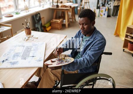 Giovane afroamericano con disabilità che mescola le vernici sulla tavolozza mentre si siede su sedia a rotelle in studio di arte da tavolo con tela Foto Stock