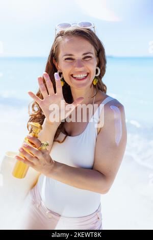 donna moderna sorridente alla spiaggia in un abbigliamento da spiaggia bianco con crema solare. Foto Stock