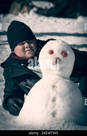 ragazzo sdraiato nella neve accanto a un pupazzo di neve autofatto con naso di carota e occhi di pomodoro, Foto Stock