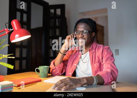 Un uomo afro-americano sorridente in uscita che parla sul telefono cellulare e chiama si siede alla scrivania in camera Foto Stock