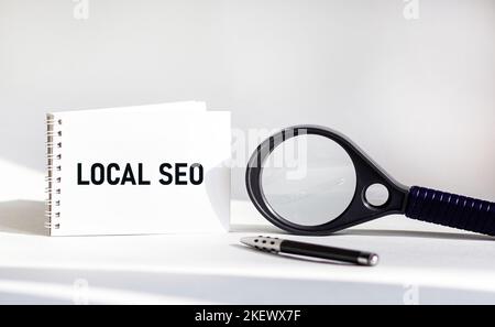 SEO locale scritto su un blocco note, su uno sfondo bianco, accanto a una lente di ingrandimento Foto Stock
