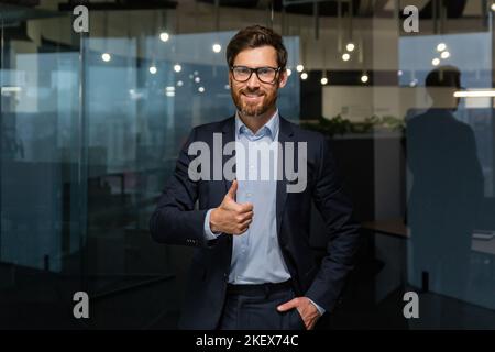 Ritratto di successo uomo d'affari maturo boss, manager in tuta d'affari occhiali e barba guardando la macchina fotografica e sorridendo in piedi vicino alla finestra, mostrando pollici in su, segno di successo e raggiungere gli obiettivi. Foto Stock