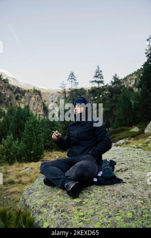 Ragazzo con cappotto e cappello che fuma una sigaretta di marijuana in natura Foto Stock